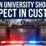 A news graphic with the text BROWN UNIVERSITY SHOOTING SUSPECT IN CUSTODY over a blurred image of the university gate and police lights